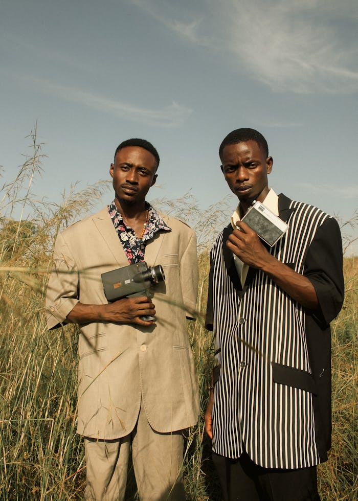 Home Two fashionable young men with vintage cameras pose confidently in a field, setting a retro yet modern vibe.