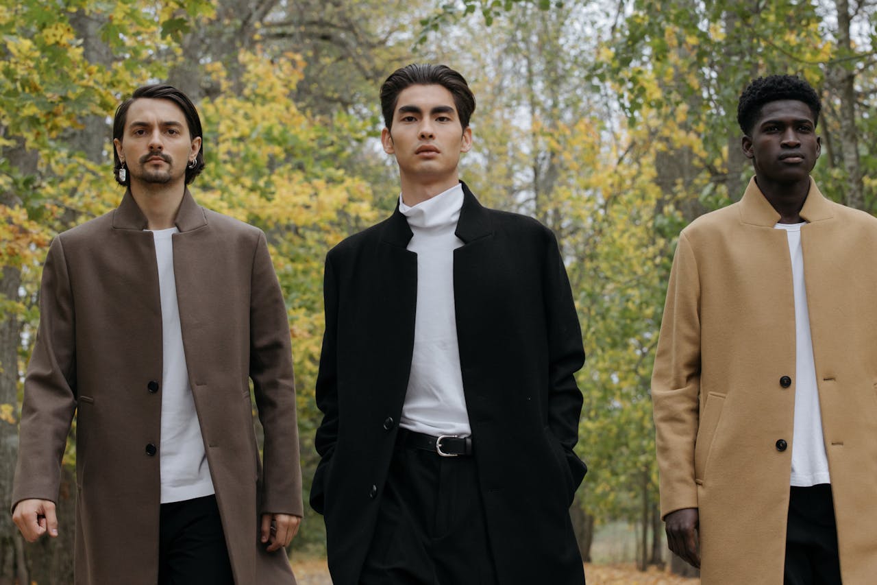 Home Three men in trendy coats walk confidently through an autumnal park, showcasing contemporary fashion.
